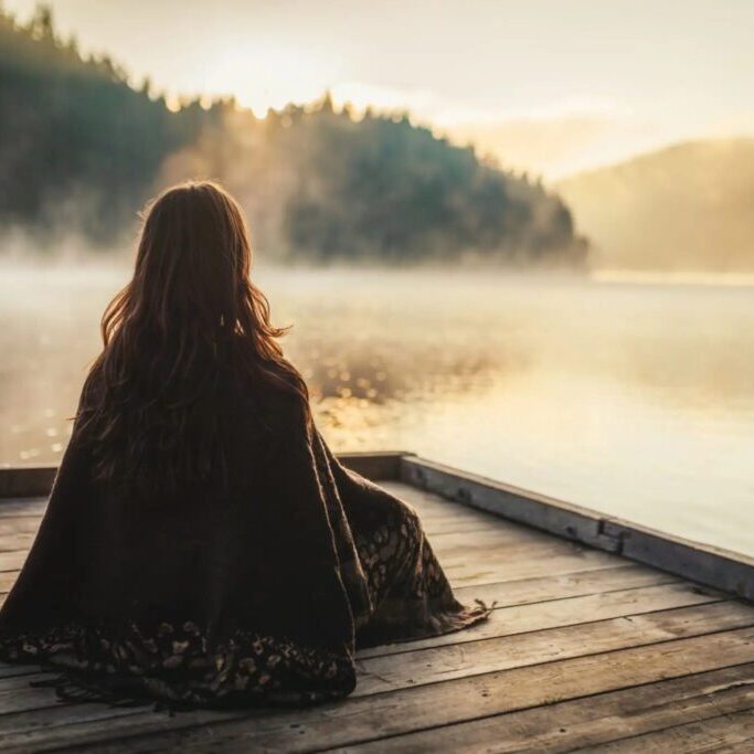 A person sitting on a dock overlooking a misty lake at sunrise.